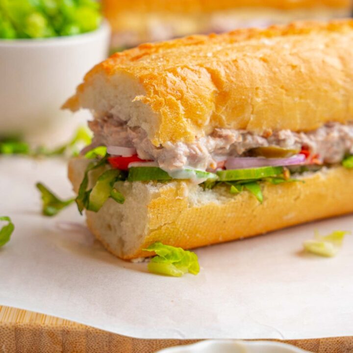 One half of a copycat Subway tuna salad sandwich served on a wooden board lined with parchment paper. The other half of the sandwich is also on the board with a bowl of shredded lettuce. The board is garnished with shredded lettuce and served on a marble counter.