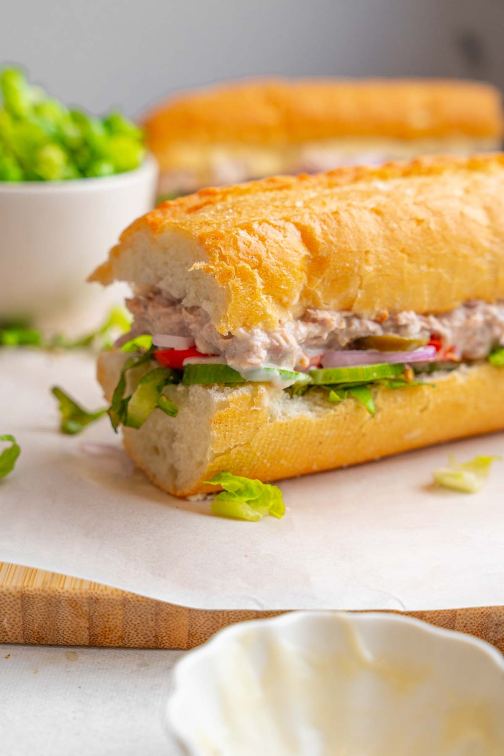 One half of a copycat Subway tuna salad sandwich served on a wooden board lined with parchment paper. The other half of the sandwich is also on the board with a bowl of shredded lettuce. The board is garnished with shredded lettuce and served on a marble counter.