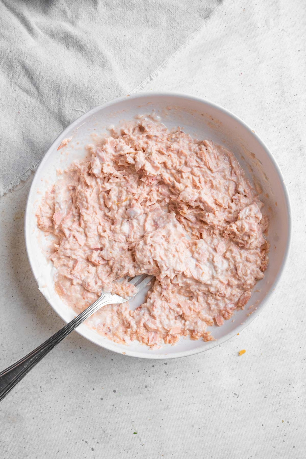 A white bowl with a spoon mixing tuna and mayonnaise. The bowl is on a marble counter with a white cloth napkin off to the side.