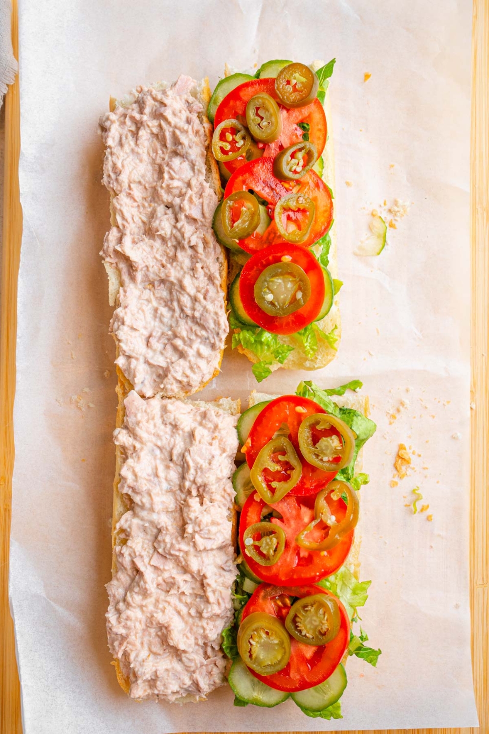 A whole copycat Subway tuna salad sandwich opened on a wooden board lined with parchment paper. The top portions of the sandwich have tuna salad while the bottom portions are layered with shredded lettuce, sliced cucumber, sliced tomato, and sliced jalapeno.