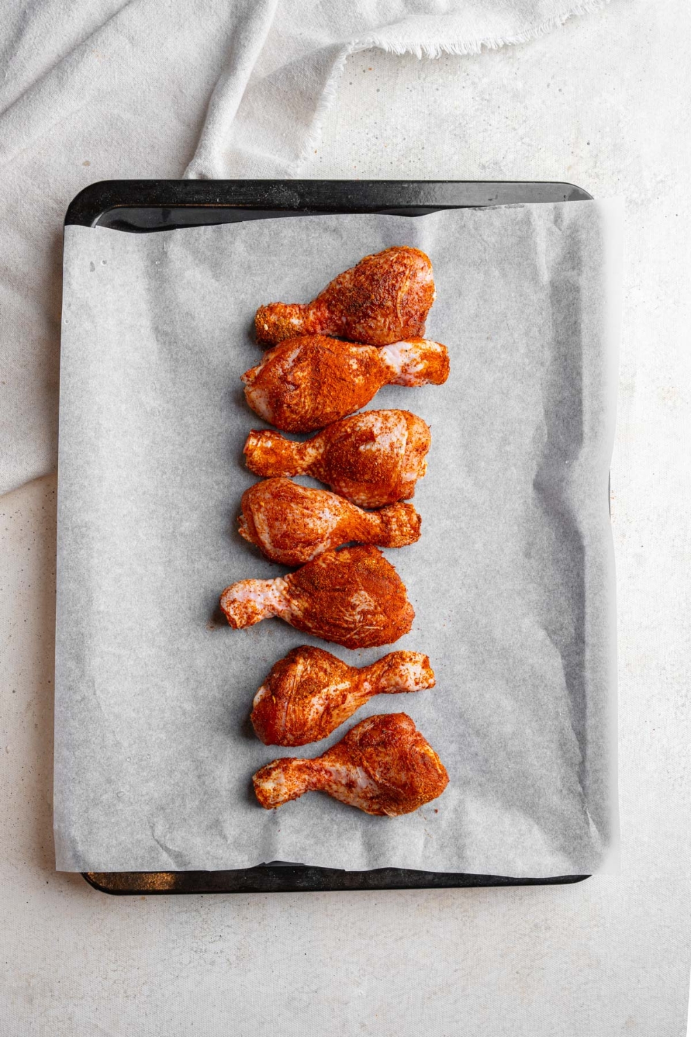 A baking sheet lined with parchment paper with seven uncooked chicken legs seaoned with various spices. The sheet is on a white counter with a white cloth napkin.