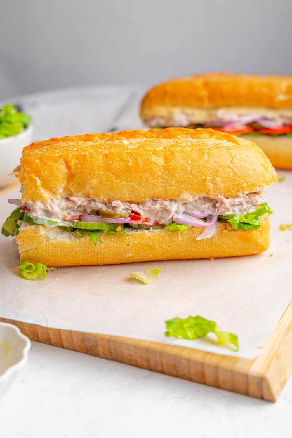 Two halves of a copycat Subway tuna sandwich served on a wooden board lined with parchment paper. The board is garnished with shredded lettuce with a bowl of shredded lettuce next to the sandwich.