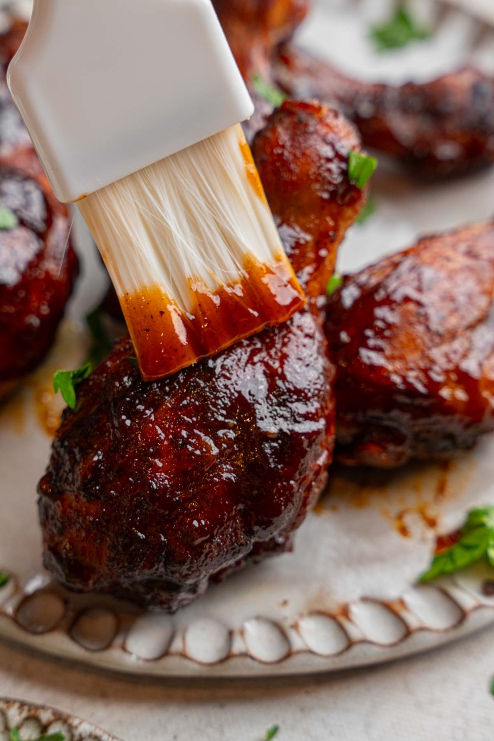 A close up of a brush brushing BBQ sauce on an oven roasted BBQ chicken leg. The chicken is on a ceramic plate with additional pieces of chicken.