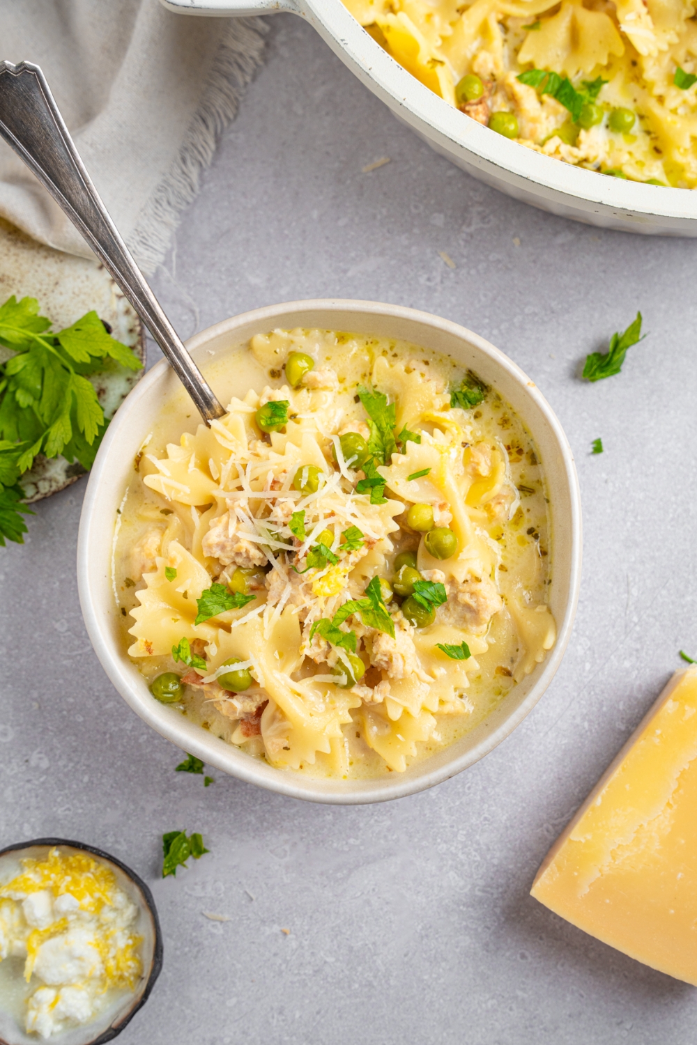 A white bowl with ground chicken soup garnished with fresh parsley and shredded parmesan cheese. There is a spoon in the soup. The bowl is on a white counter with a large dutch oven of soup and garnishes.