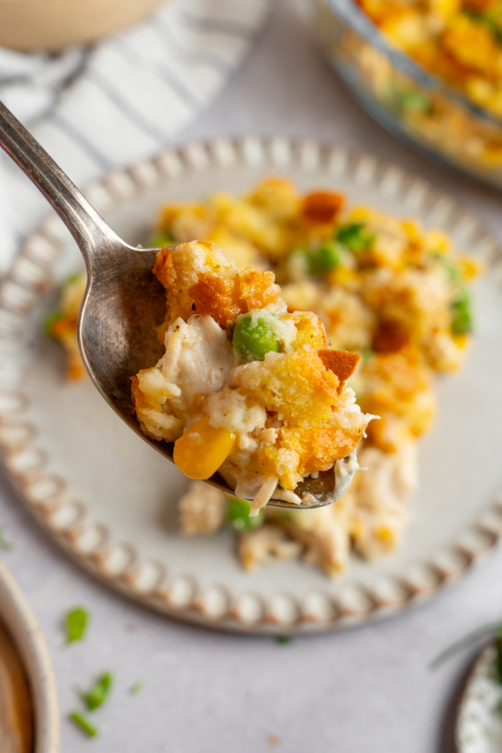 A close up of a spoon with a bite of rrotisserie chicken and stuffing casserole. There is a ceramic plate with casserole on a white counter blurred in the background.