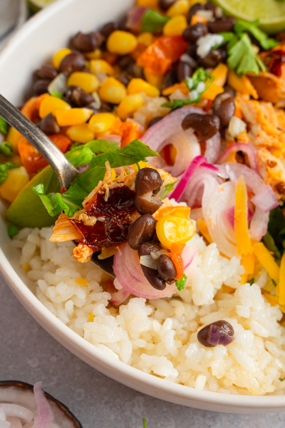 A fork taking a bite from a rotisserie chicken burrito bowl including rice, beans, corn, pickled red onion, and chicken. The bowl is garnished with fresh cilantro.