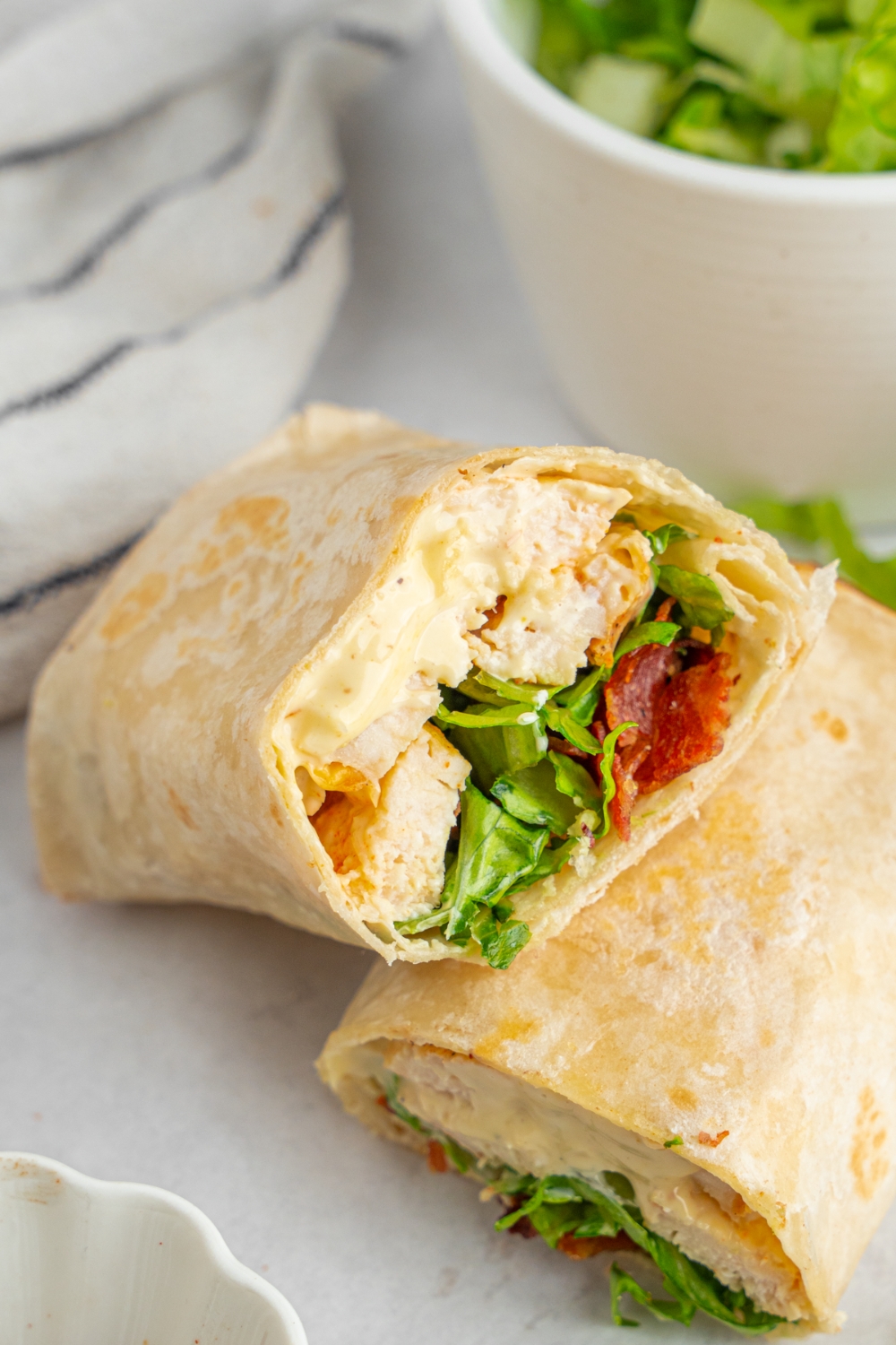 A rotisserie chicken caesar wrap sliced in half on a white counter. There is a white striped napkin and bowl of shredded lettuce.