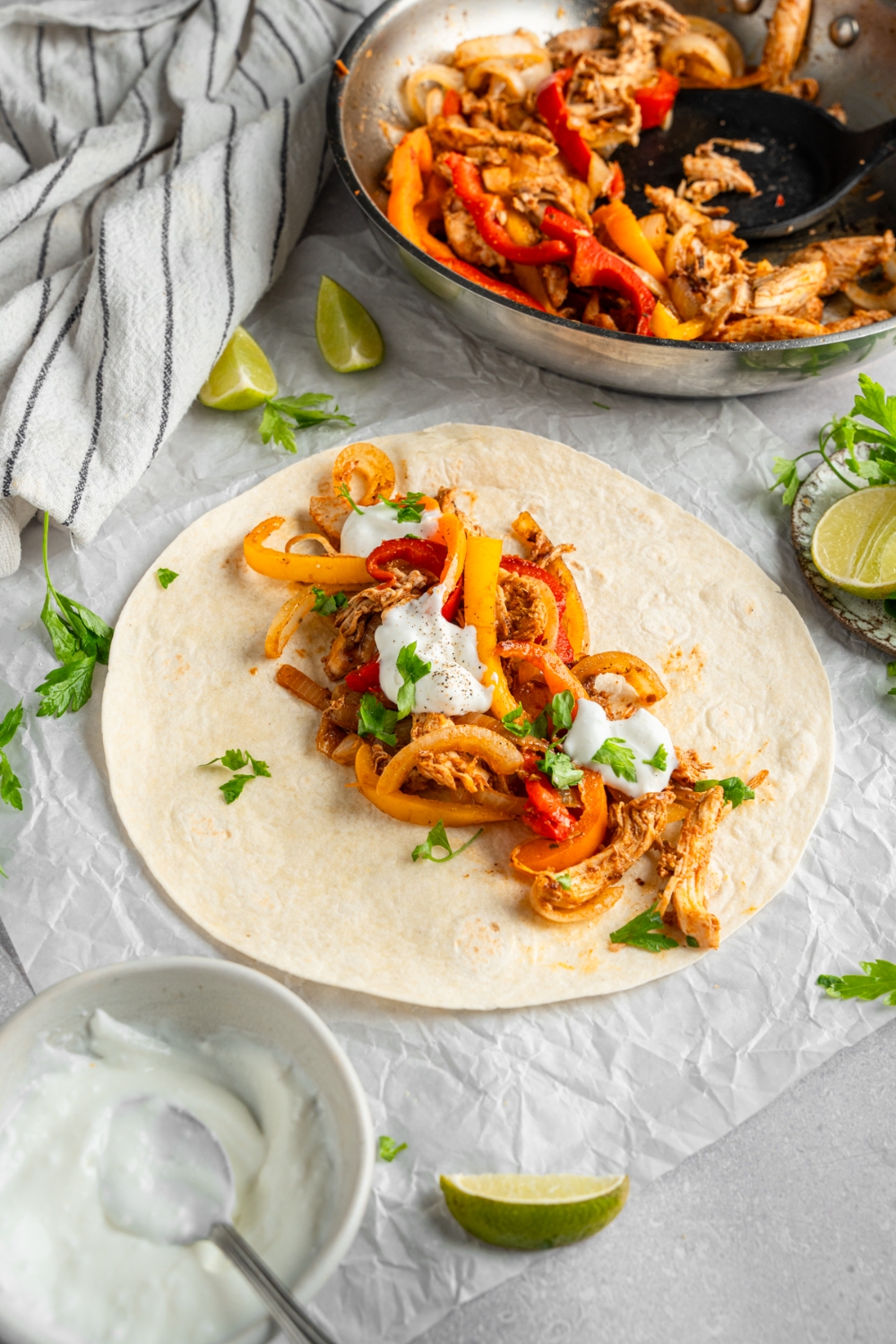 An open rotisserie chicken fajita topped with sour cream and fresh. cilantro. The fajita is on a piece of parchment paper on a white counter with a skillet of chicken fajitas and lime wedges.