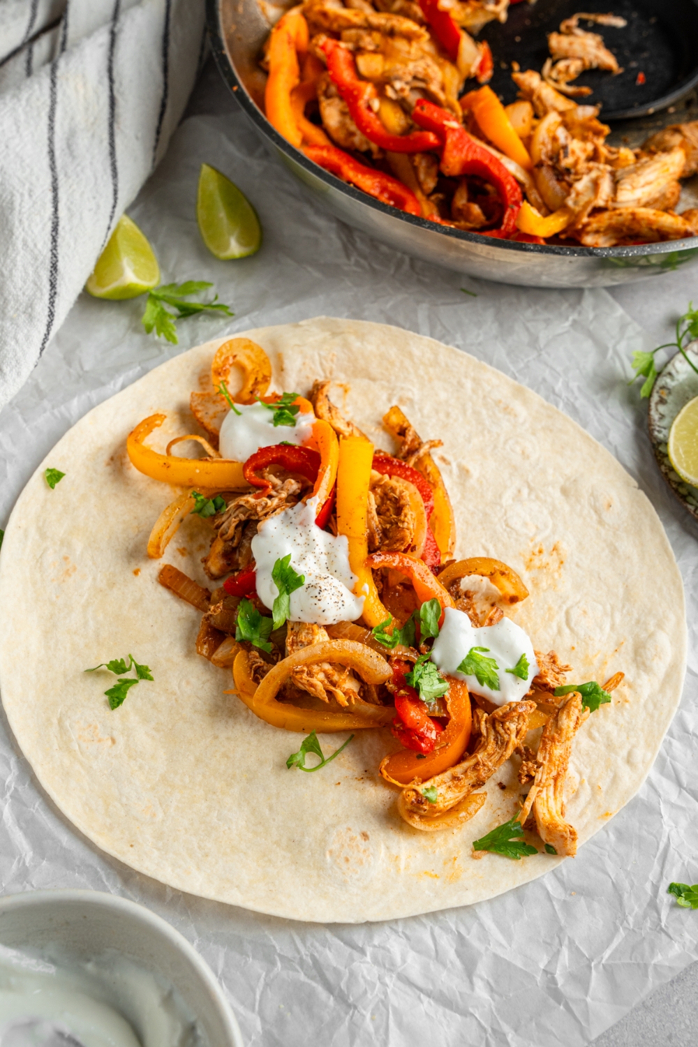 An open rotisserie chicken fajita topped with sour cream and fresh. cilantro. The fajita is on a piece of parchment paper on a white counter with a skillet of chicken fajitas and lime wedges.