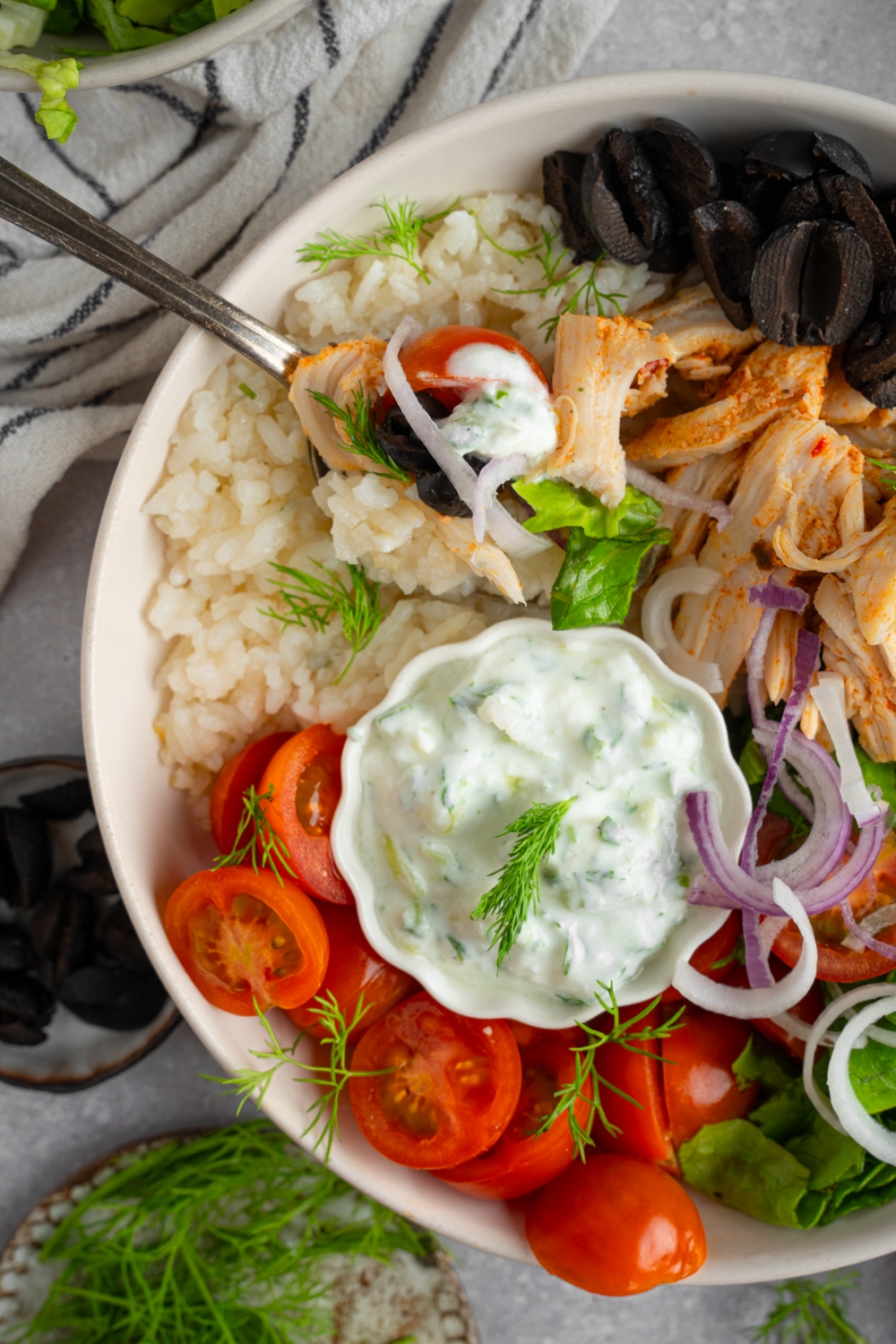 Rotisserie chicken greek bowl served in a white bowl with lettuce, rotisserie chicken, rice, black olives, tomatoes, red onion, dill, and a small bowl of tzatziki. There is a fork in the bowl.