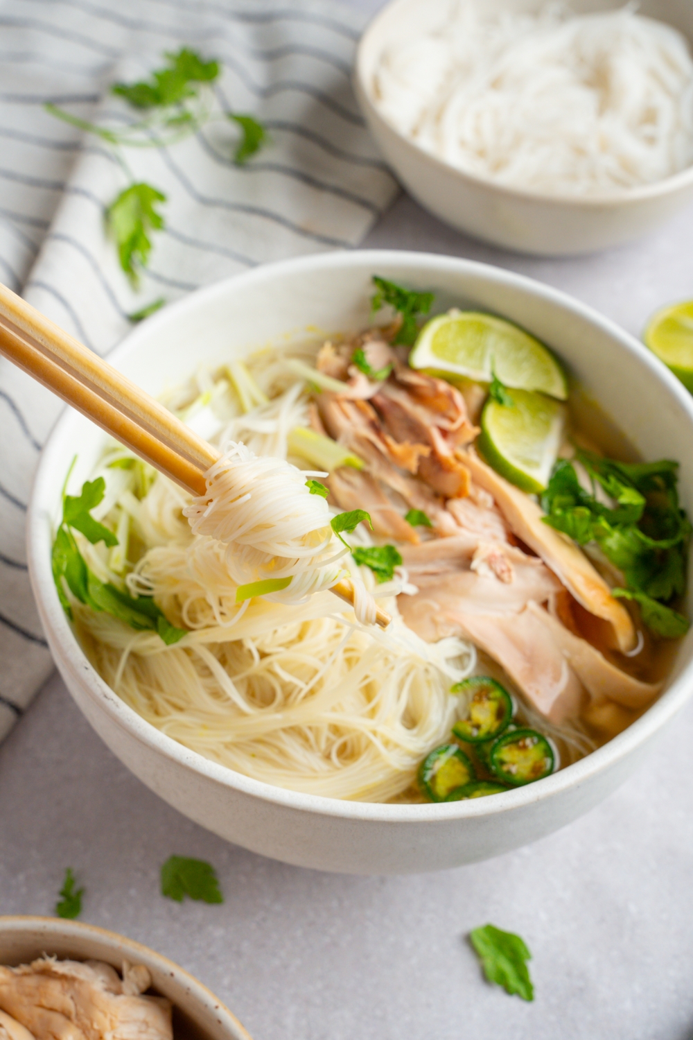 Rotisserie chicken pho served in a white bowl garnished with lime, fresh basil leaves, cilantro, bean sprouts, sliced jalapeno, and green onions. A pair of chopsticks is taking a bite of pho noodles. The bowl is on a white counter with a white bowl of cooked pho noodles and a white striped napkin.