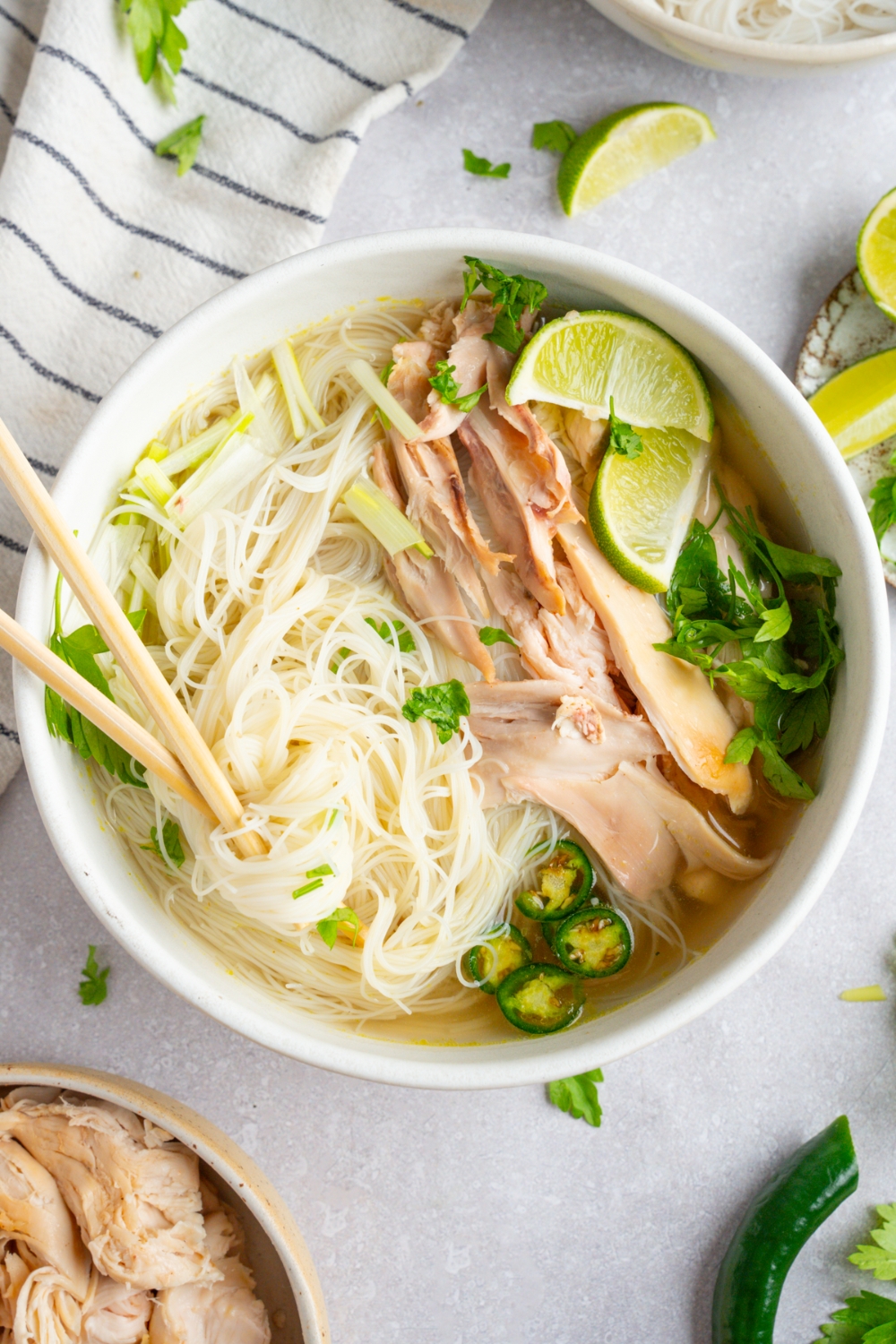 Rotisserie chicken pho served in a white bowl garnished with lime, fresh basil leaves, cilantro, bean sprouts, sliced jalapeno, and green onions. A pair of chopsticks is in the bowl. The bowl is on a white counter with a bowl of rotisserie chicken and a white striped napkin.