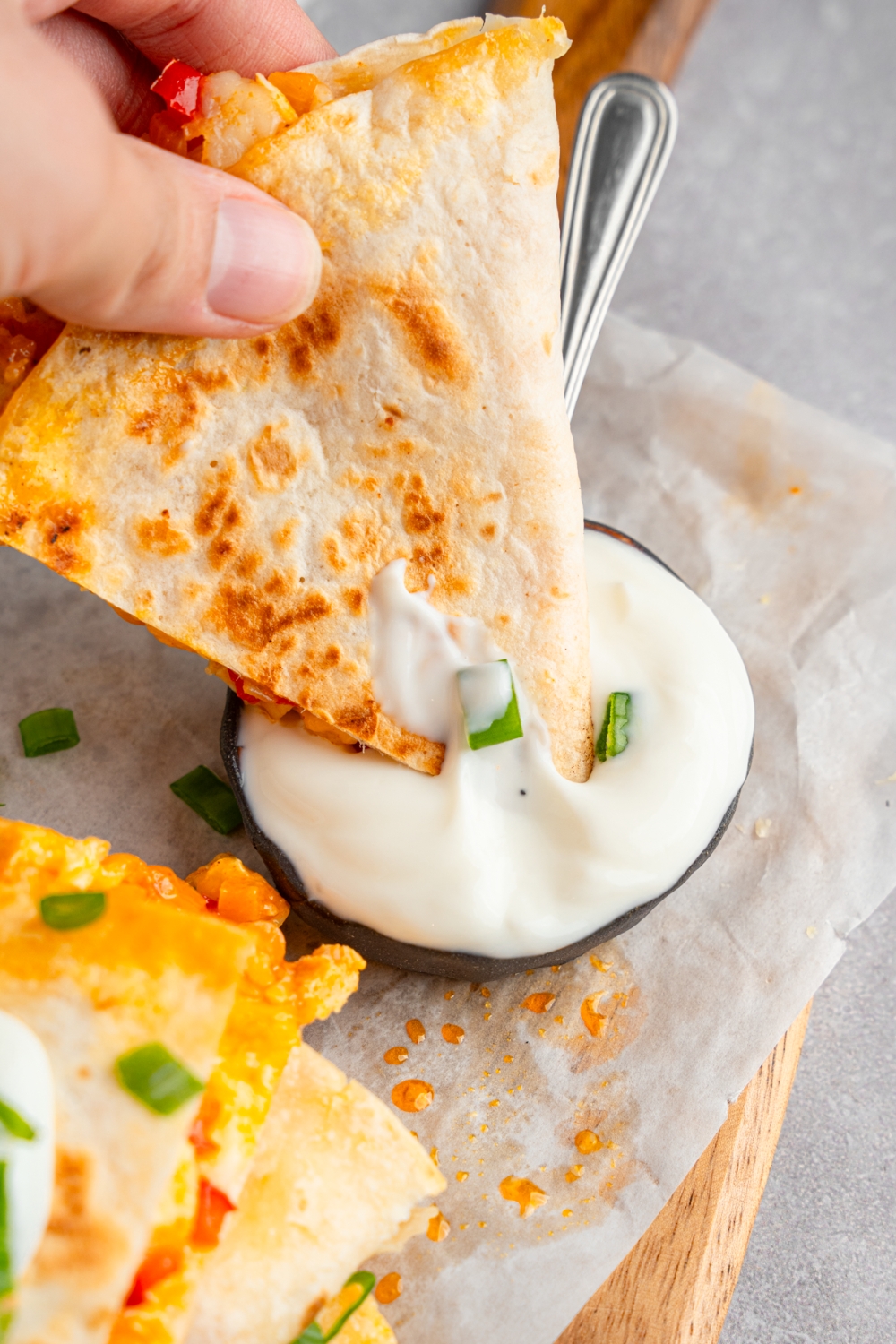 A hand dipping a slice of rotisserie chicken quesadillas into a cup of sour cream with a spoon. The sauce is on a wooden board lined with parchment paper with additional slices of quesadilla garnished with sliced green onion and lime.