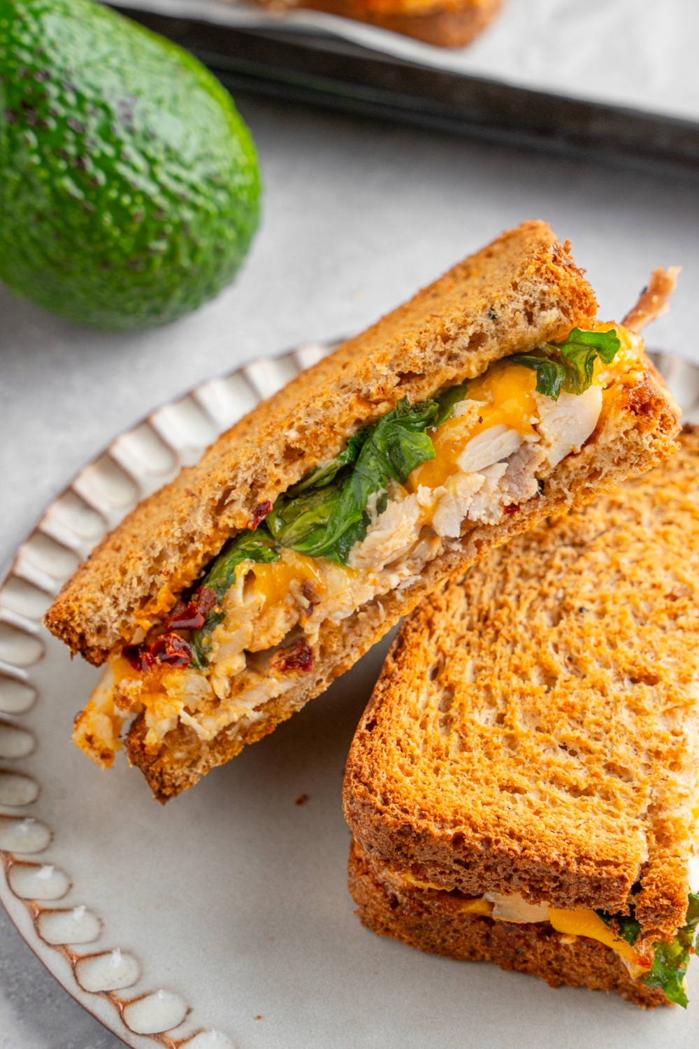 A rotisserie chicken sandwich with melted cheese, spinach, and chipotle mayo sliced in half and served on a ceramic plate. The plate is on a white counter with an avocado.