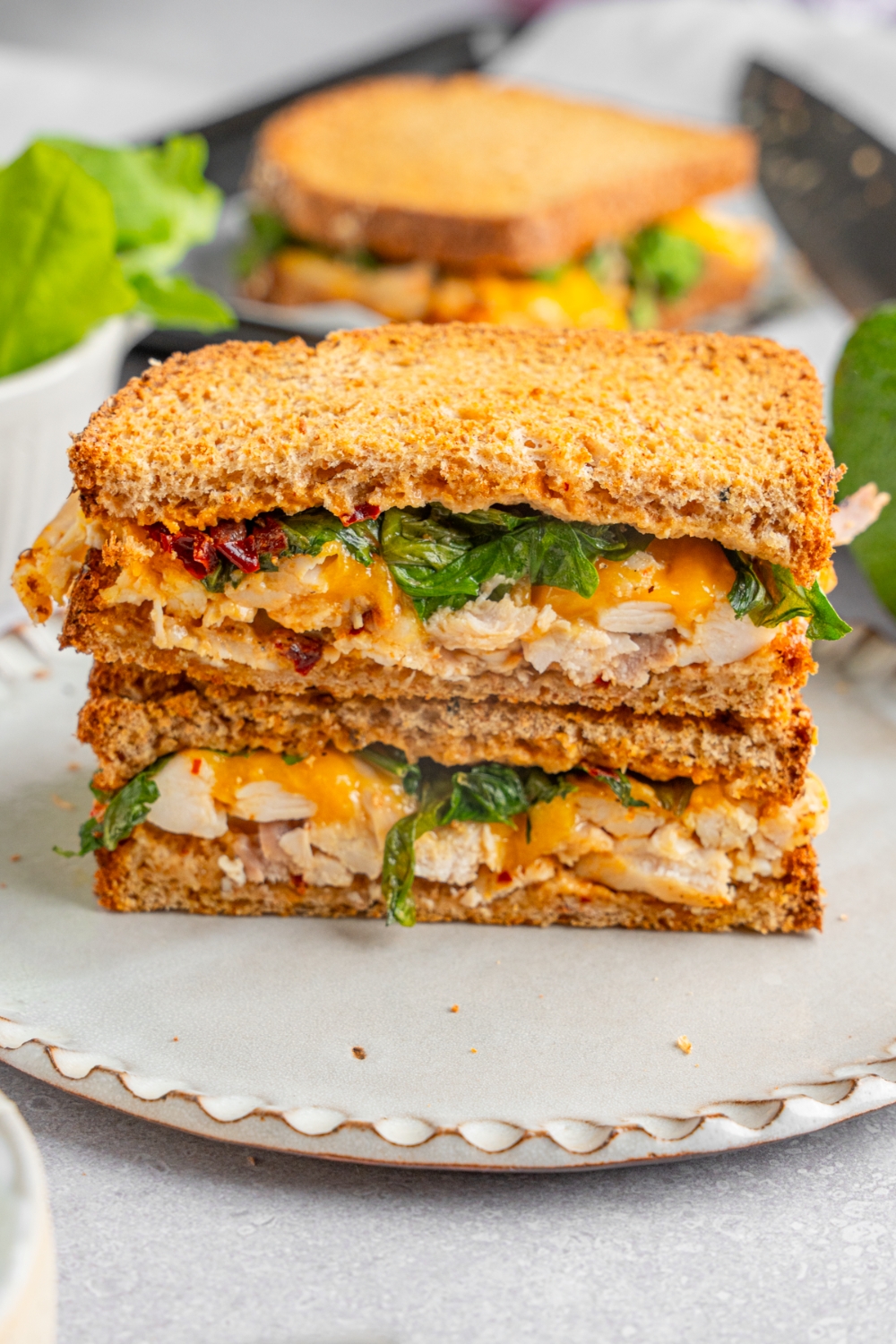 A rotisserie chicken sandwich with chipotle mayo, spinach, and melted cheese sliced in half with each half stacked on top of the other. The sandwich is on a ceramic plate. The plate is on a white counter with an additional sandwich in the background.