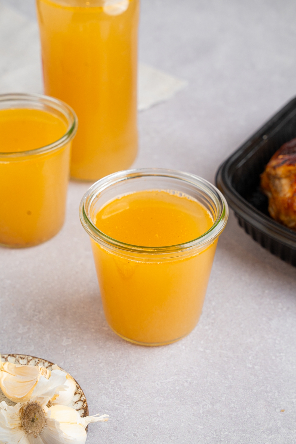 A glass jar with rotisserie chicken bone broth. The jar is on a white counter with a second jar and pitcher of bone broth. There is a tray of rotisserie chicken next to the jars.