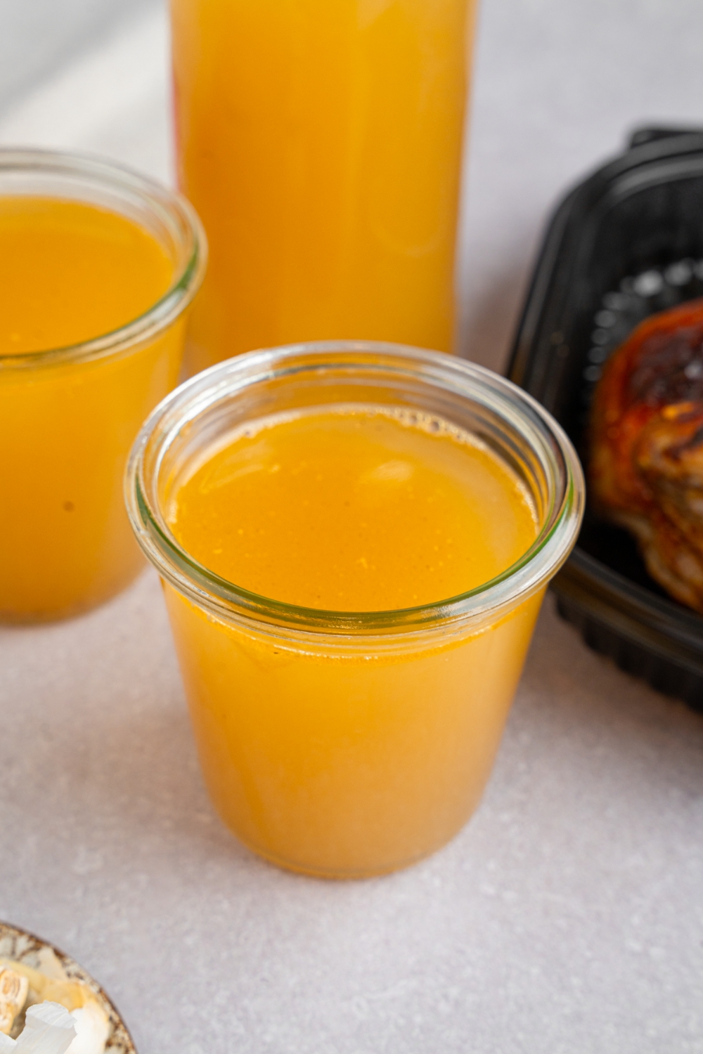 A glass jar with rotisserie chicken bone broth. The jar is on a white counter with a second jar and pitcher of bone broth. There is a tray of rotisserie chicken next to the jars.