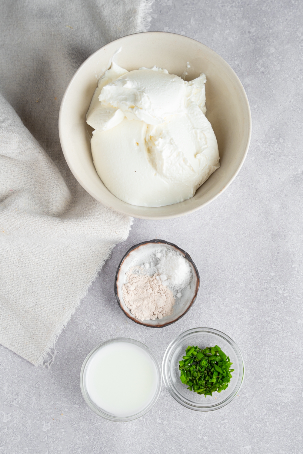 An overhead shot of four bowls in various sizes containing ingredients for chive and onion cream cheese including cream cheese, milk, onion powder, chopped chives, and salt.