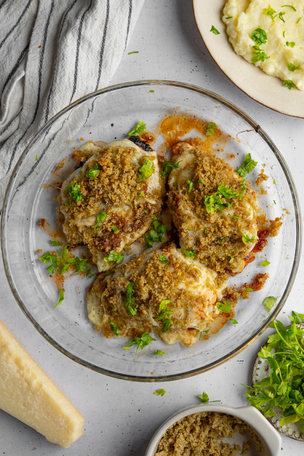 A glass baking dish with three pieces of copycat Longhorn parmesan crusted chicken garnished with fresh parsley. The dish is on a white counter with a plate of mashed potatoes and garnishes.