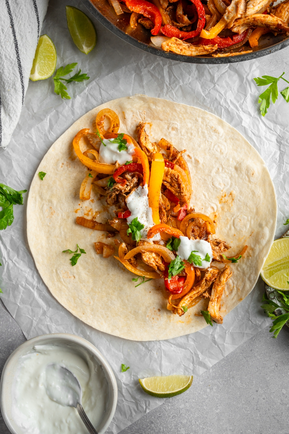 An open rotisserie chicken fajita topped with sour cream and fresh. cilantro. The fajita is on a piece of parchment paper on a white counter with a skillet of chicken fajitas and lime wedges.