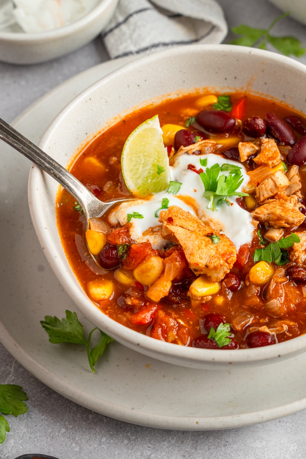 A spoon in a bowl of rotisserie chicken taco soup. The soup is in a white bowl topped with a dollop of sour cream, lime slice, and fresh cilantro. The bowl is on a white plate.