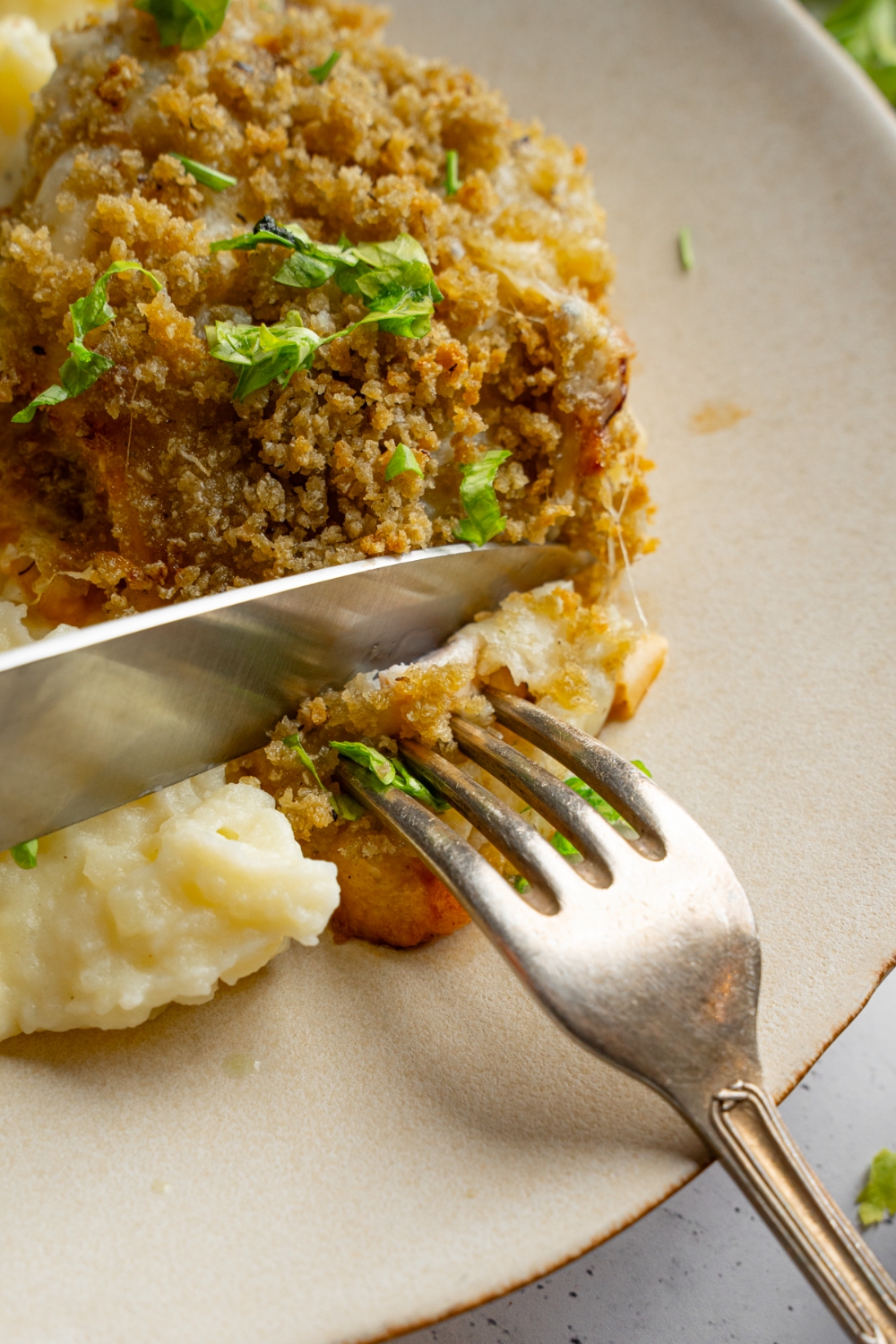 A fork and knife slicing a bite of copycat Longhorn parmesan crusted chicken served with mashed potatoes.