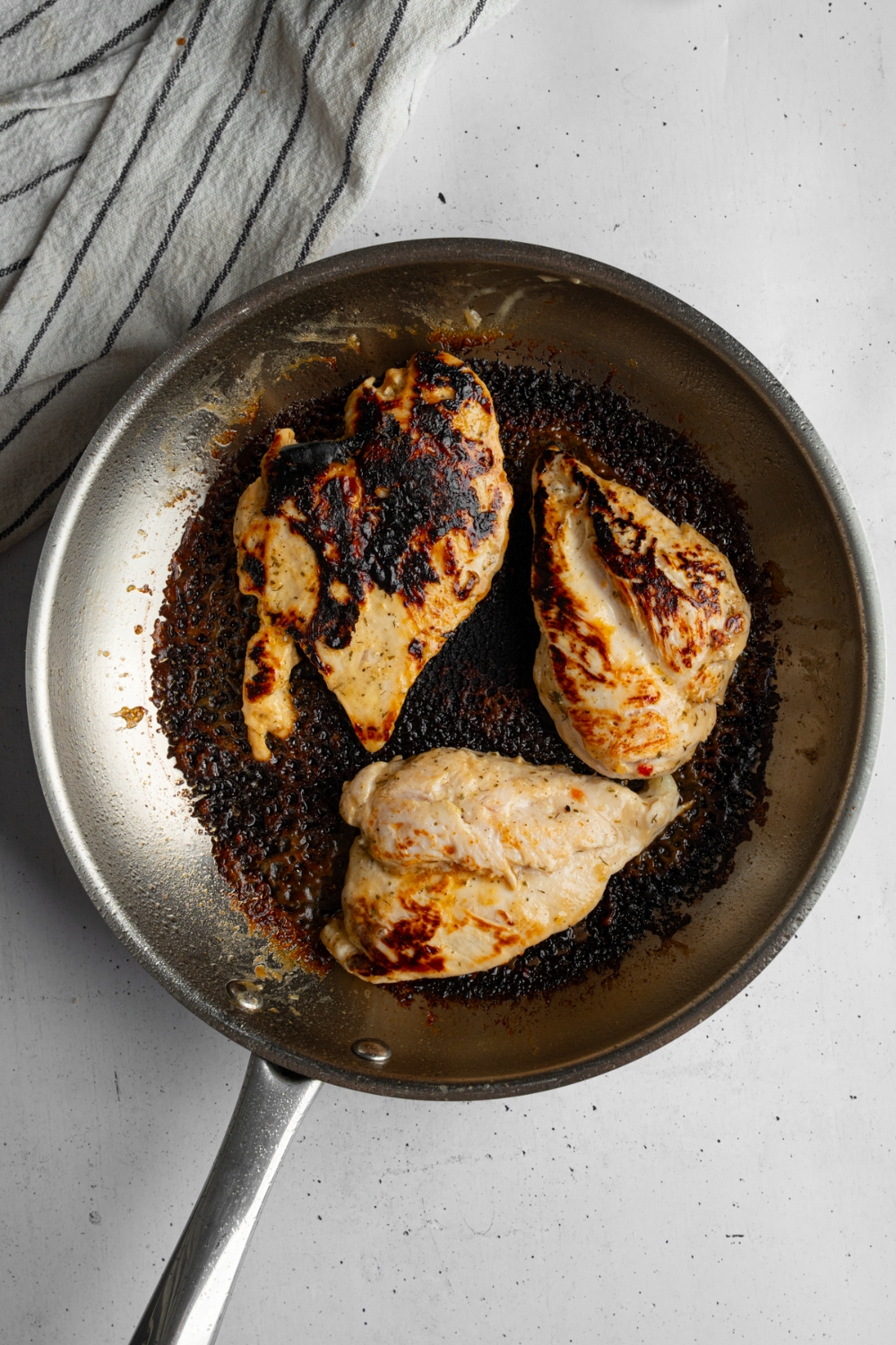 A skillet with three chicken breasts cooking in butter. The skillet is on a white counter with a white cloth napkin.