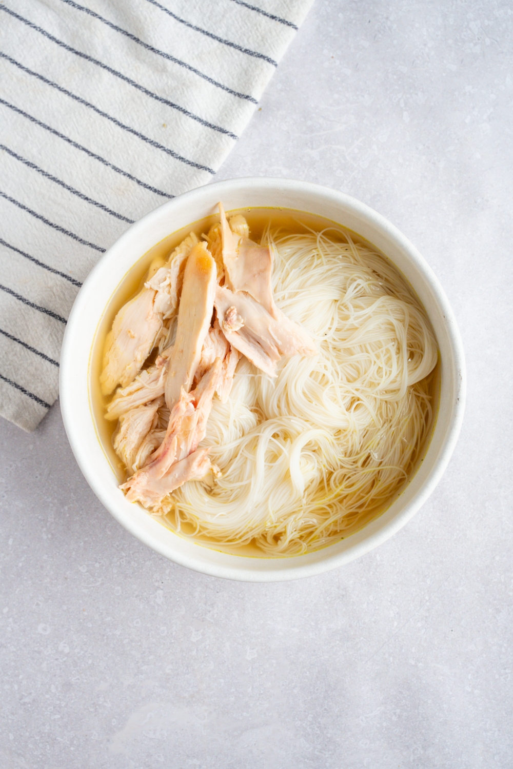 A white bowl with cooked pho noodles, rotisserie chicken, and pho broth. The bowl is on a white counter with a white striped napkin.
