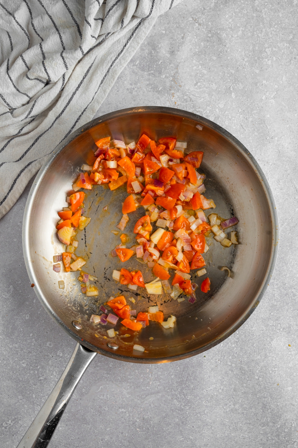 A stainless steel skillet with tomatoes and onion cooking in oil. The skillet is on a white counter with a white striped napkin.
