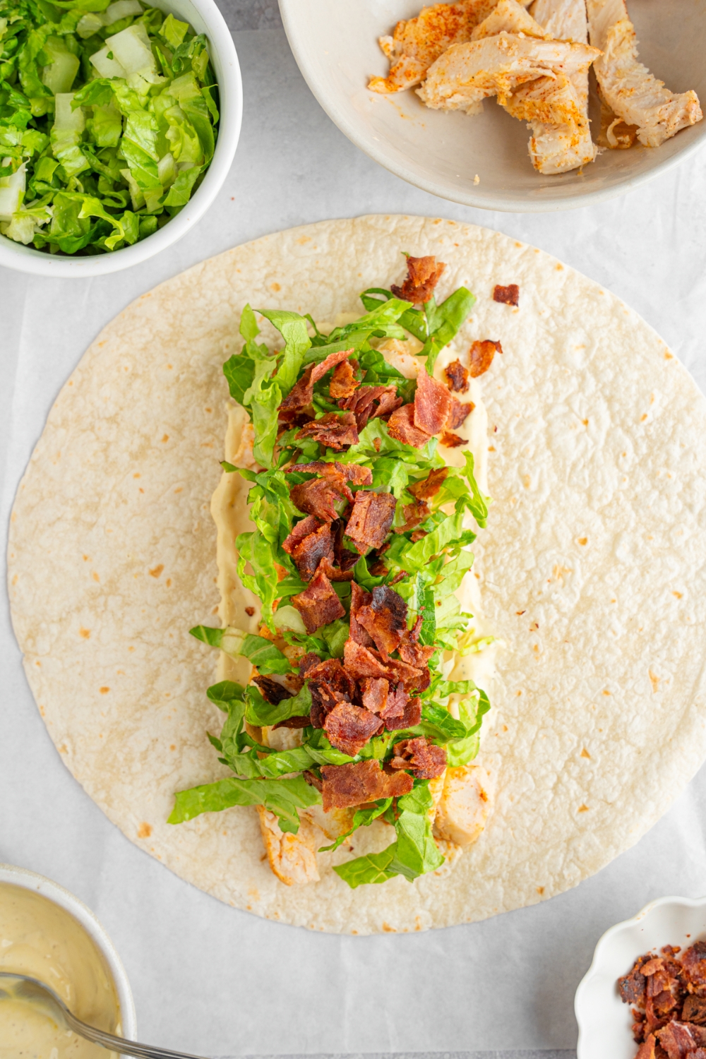 An open rotisserie chicken caesar wrap including caesar dressing, chicken, shredded lettuce, and chopped bacon. The wrap is on a white counter with a bowl of shredded lettuce and bowl of chicken.
