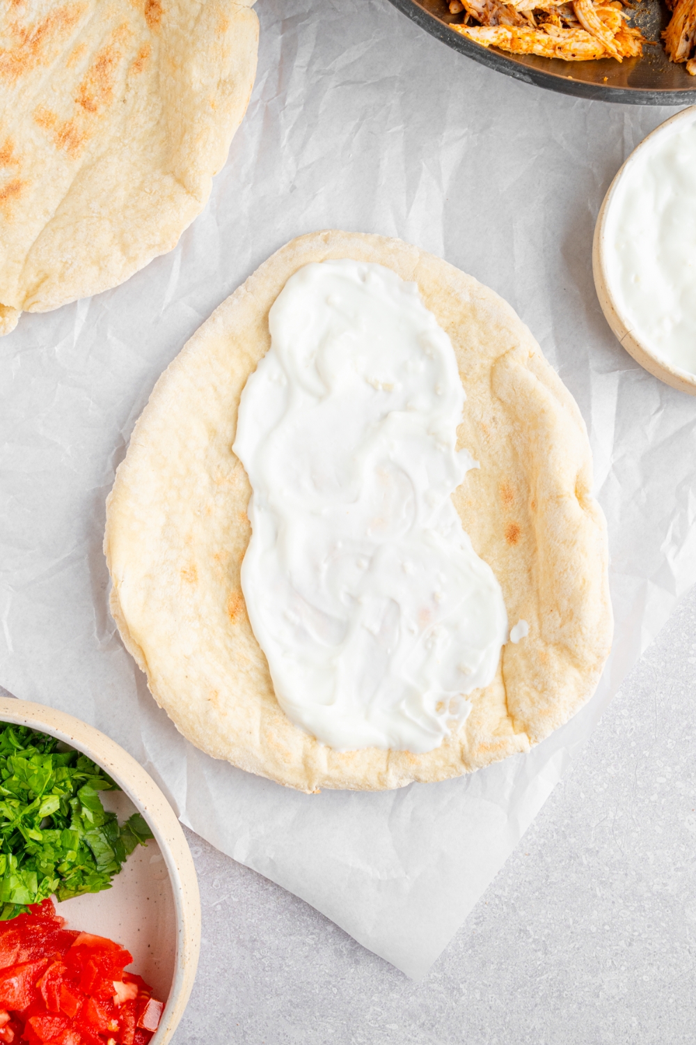 An open pita topped with garlic sauce. The pita is on a piece of parchment paper with an additional pita and small bowl of garnishes.