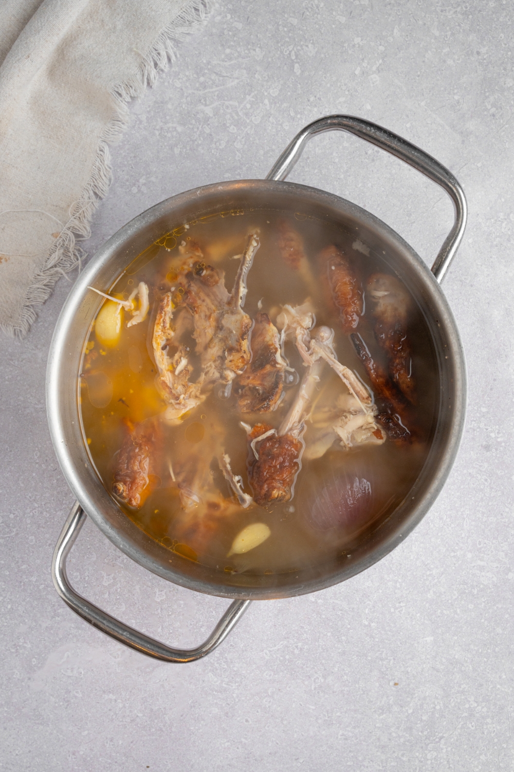A stock pot filled with ingredients cooking to make bone broth including rotisserie chicken, water, onion, and garlic. The pot is on a white counter with a white cloth napkin.