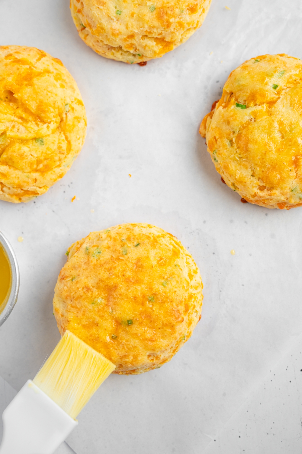 Four baked cheddar chive biscuits on parchment paper. One of the biscuits is being brushed with butter using a pastry bush.