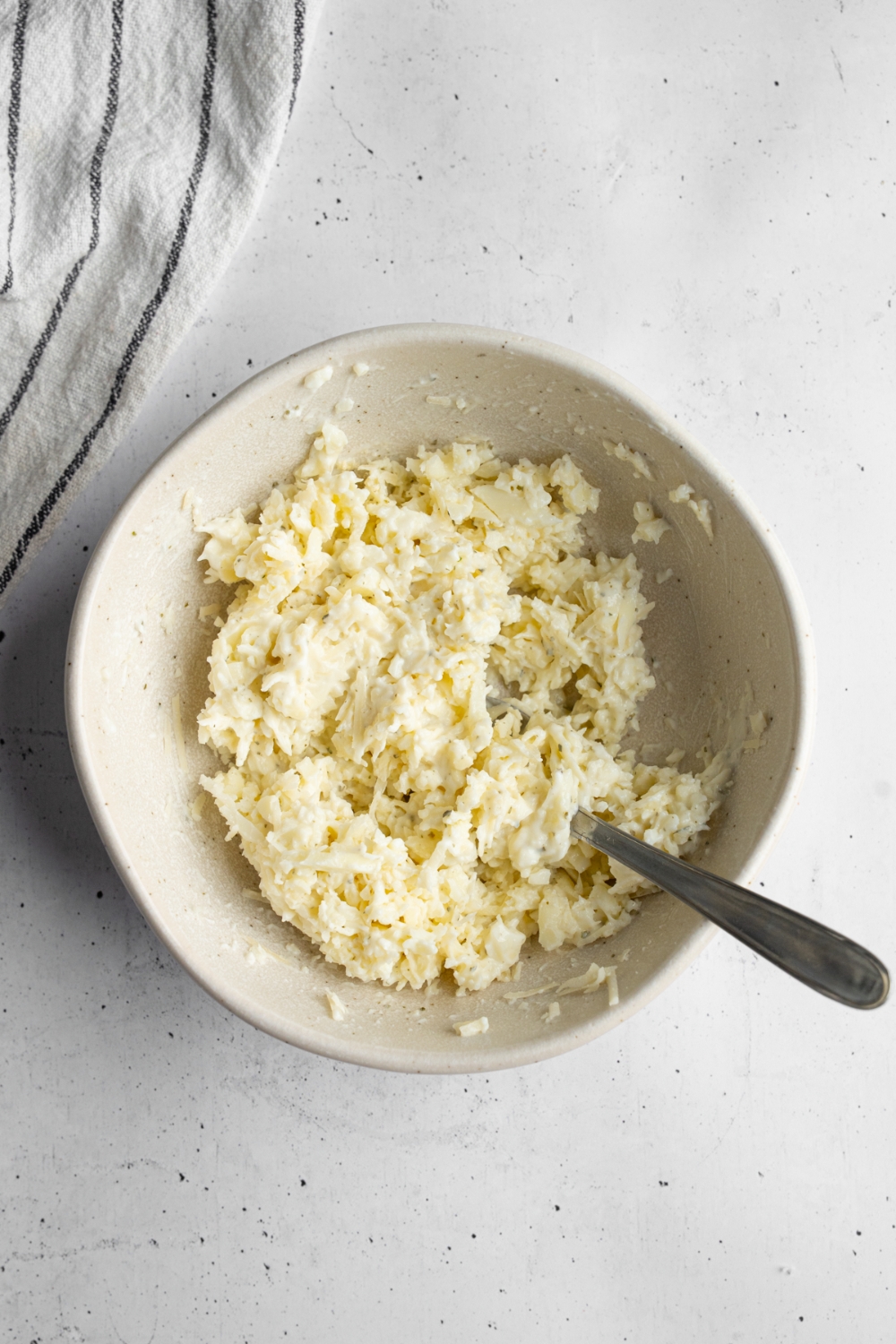A white bowl with a spoon mixing cheeses and ranch seasoning. The bowl is on a white counter with a white striped napkin.