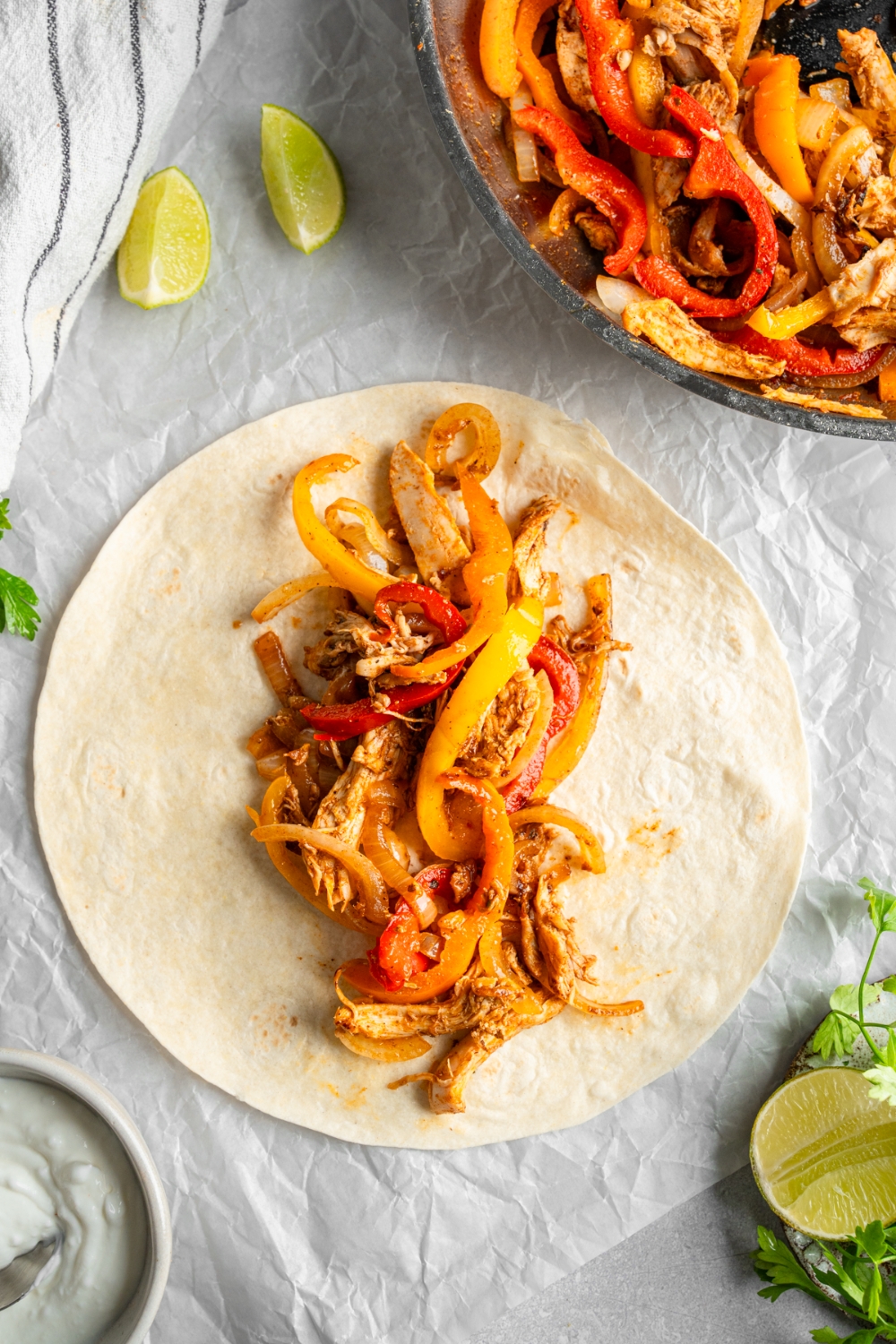 An open rotisserie chicken fajita topped with roasted bell peppers. The fajita is on a piece of parchment paper on a white counter with a skillet of chicken fajitas and lime wedges.
