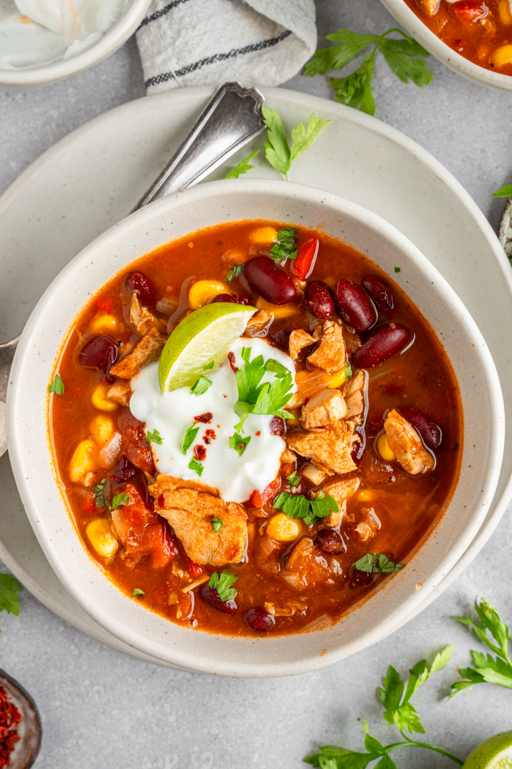 A white bowl with rotisserie chicken taco soup garnished with sour cream, lime, and fresh cilantro. The bowl is on a white ceramic plate with a spoon on a white counter with fresh cilantro.