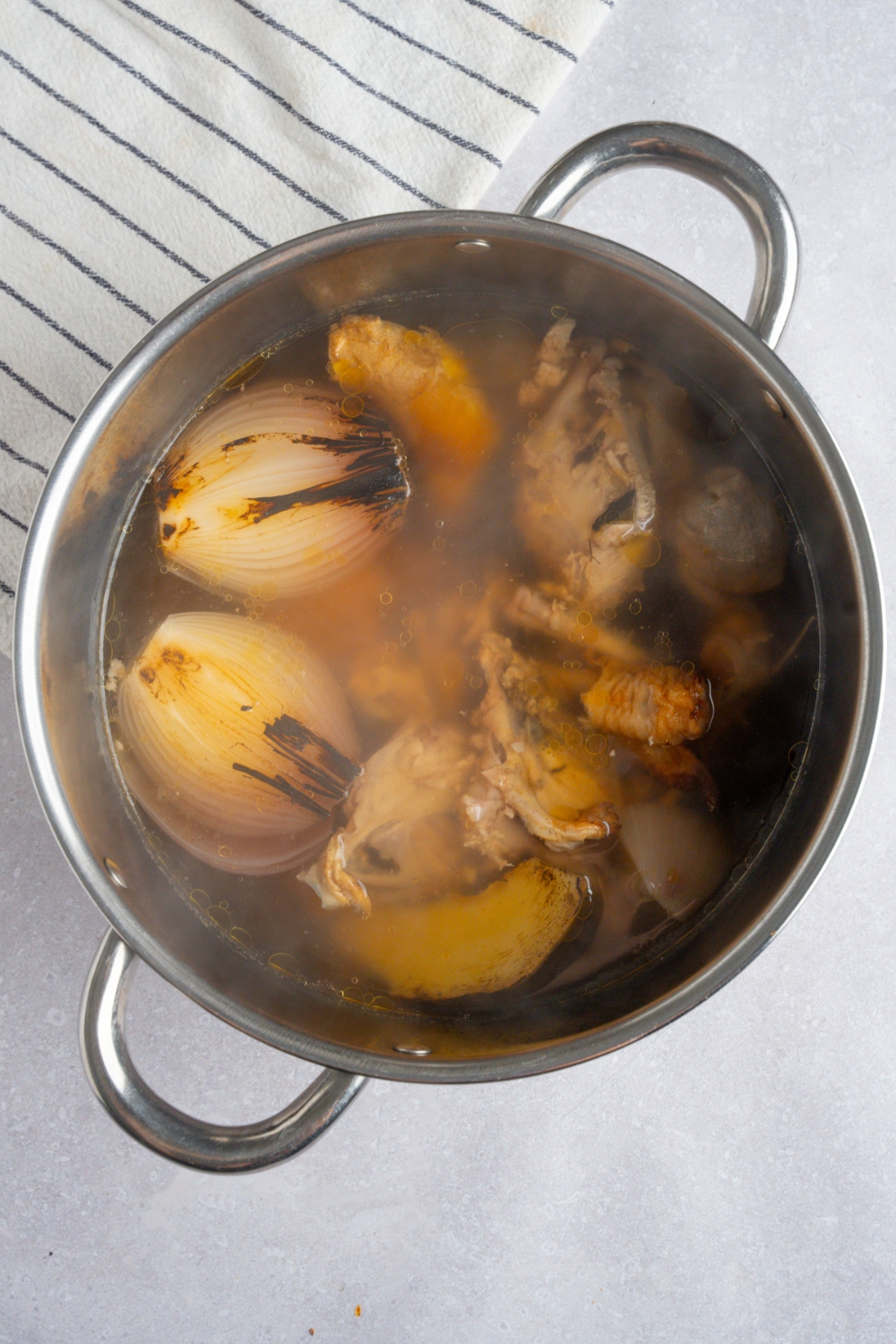 A stock pot with ingredients cooking to make rotisserie chicken pho broth. The pot is on a white counter with a white striped napkin.