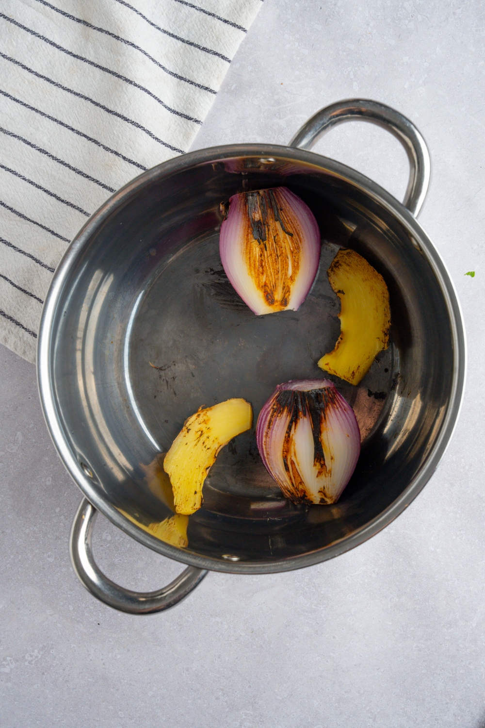 A stock pot with charred red onion and ginger. The pot is on a white counter with a white striped napkin.