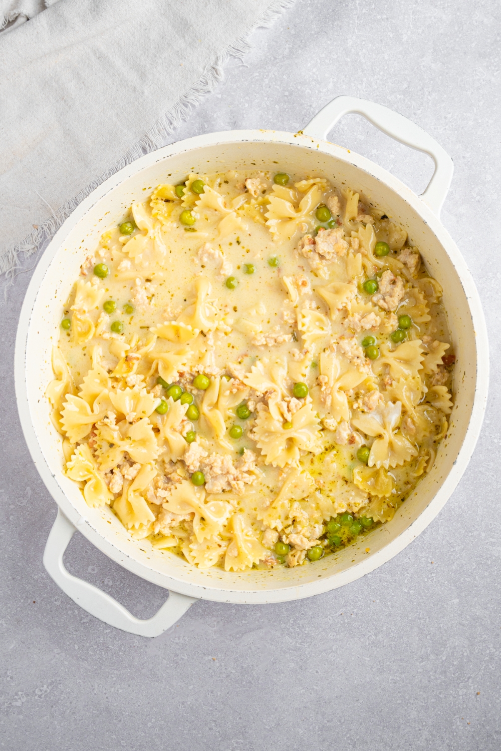 A large white pot with ground chicken soup with pasta, peas, and ricotta cream. The pot is on a white counter with a white cloth napkin.