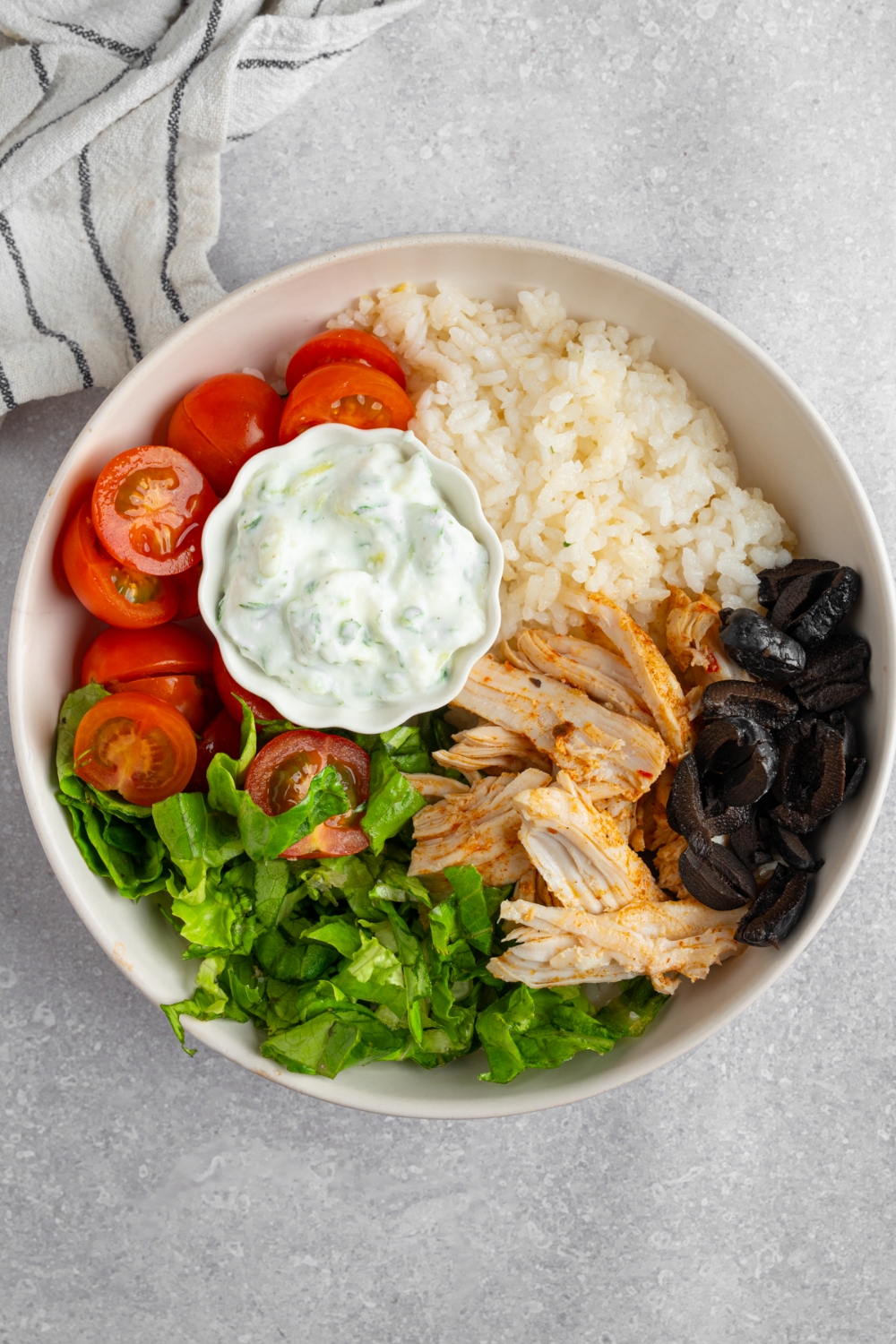 Rotisserie chicken greek bowl served in a white bowl with lettuce, rotisserie chicken, rice, black olives, tomatoes, red onion, and a small bowl of tzatziki. The bowl is on a white counter with a white striped napkin.