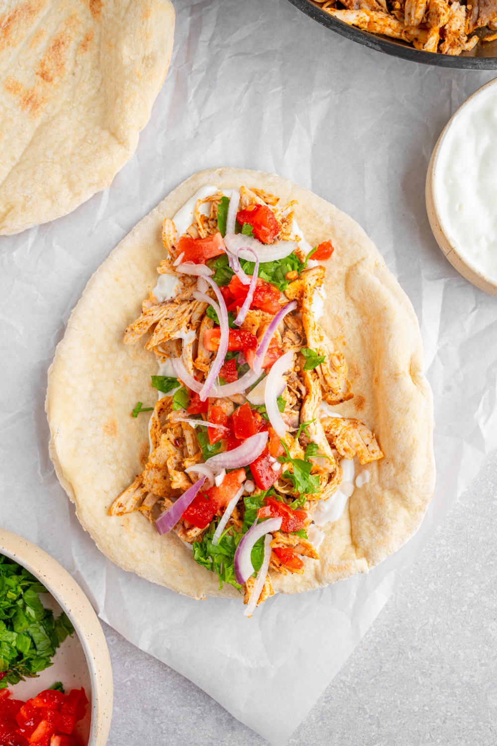 An open chicken shawarma including a pita topped with rotisserie chicken, garlic sauce, red onion, tomato, and fresh parsley. The parchment paper is on a white counter with additional pita and small bowl of garlic sauce.