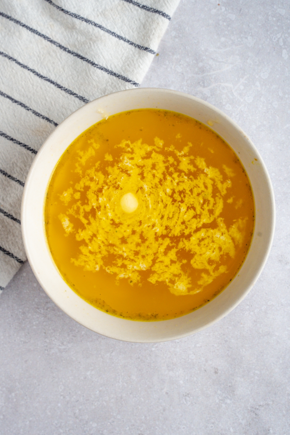 A white bowl with butter melted with chicken broth. The bowl is on a white counter with a white striped napkin.