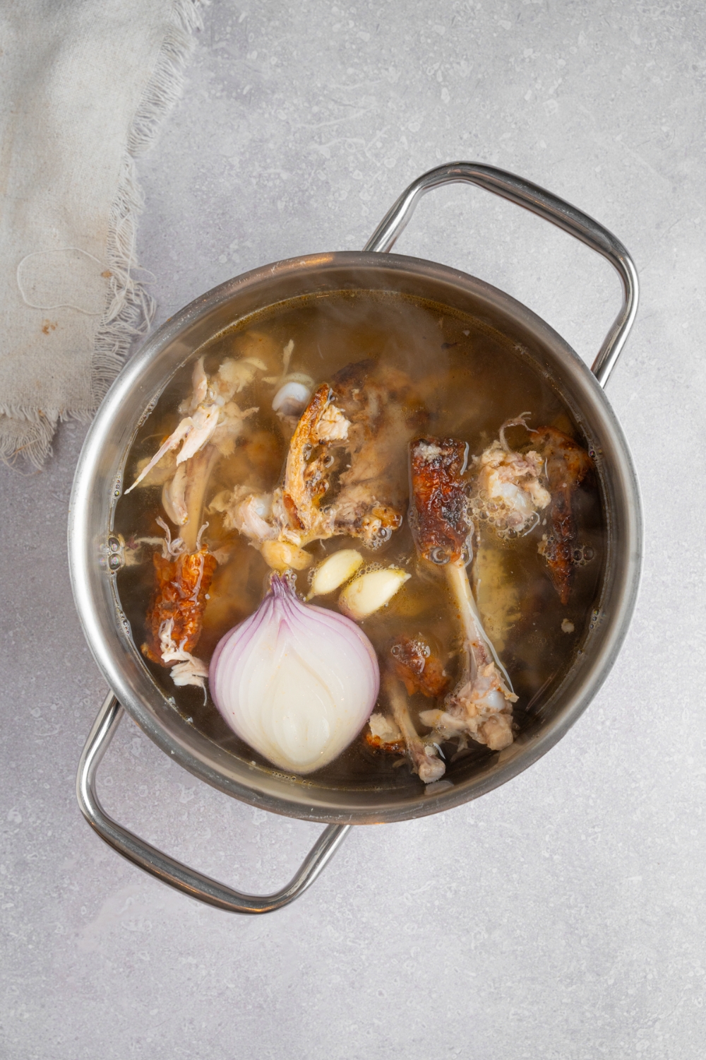 A large stock pot filled with ingredients to make bone broth including rotisserie chicken parts, water, garlic, and onion. The pot is on a white counter with a white cloth napkin.