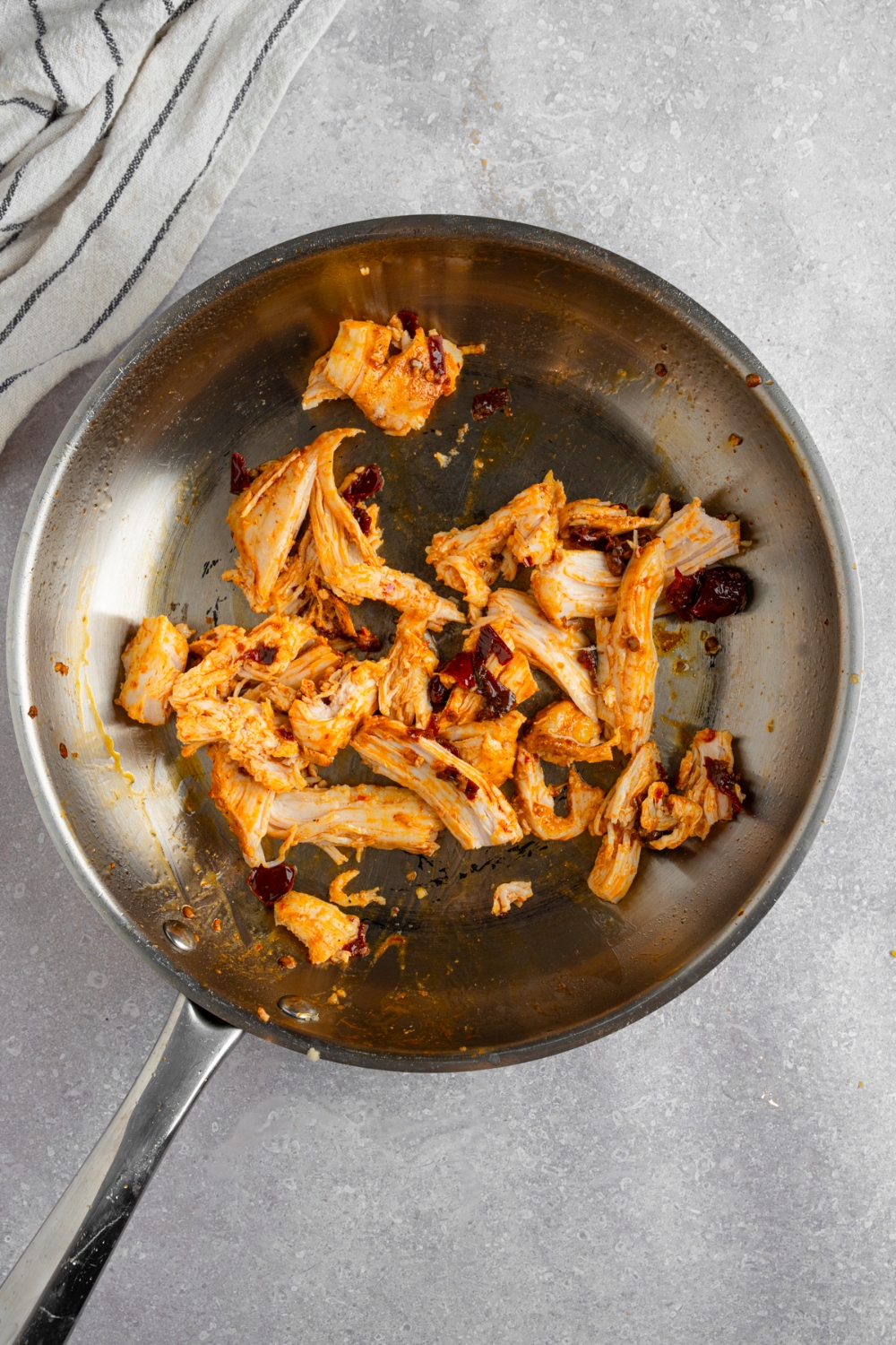 A skillet with rotisserie chicken cooking in seasonings. The skillet is on a white counter with a white striped napkin.