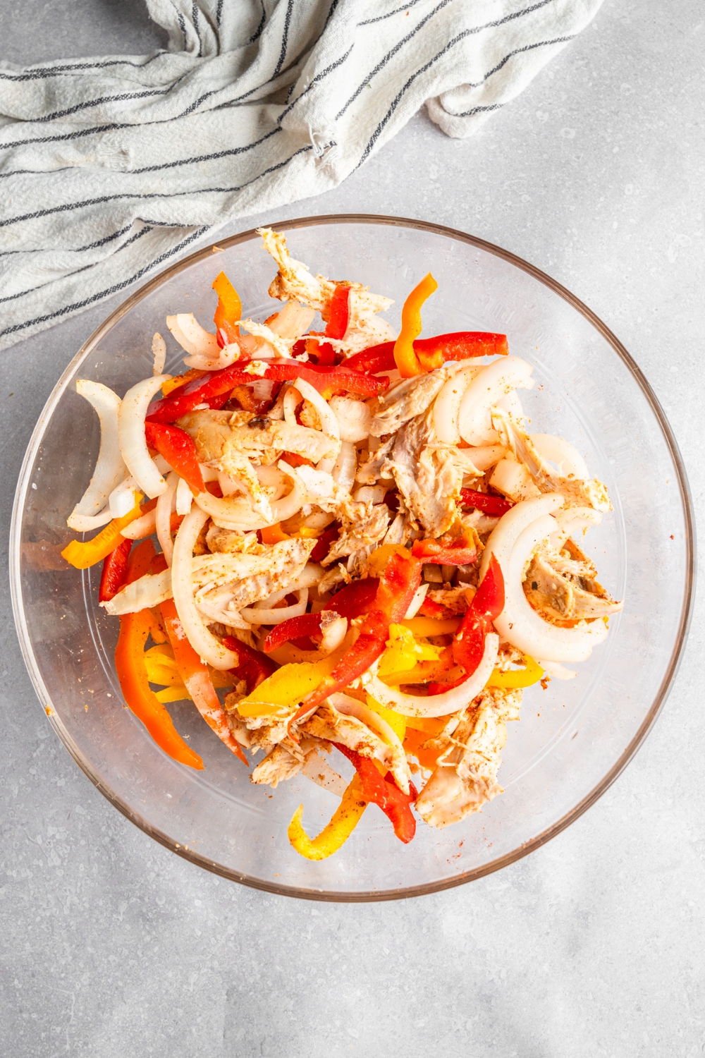 A glass bowl with shredded rotisserie chicken and sliced bell peppers. The bowl is on a white counter with a white striped napkin.