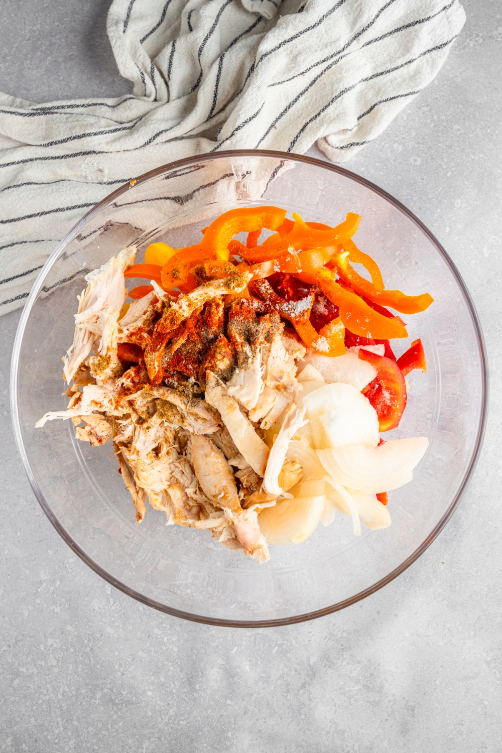 A glass bowl with shredded rotisserie chicken, sliced bell peppers, onions, and seasonings. The bowl is on a white counter with a white striped napkin.