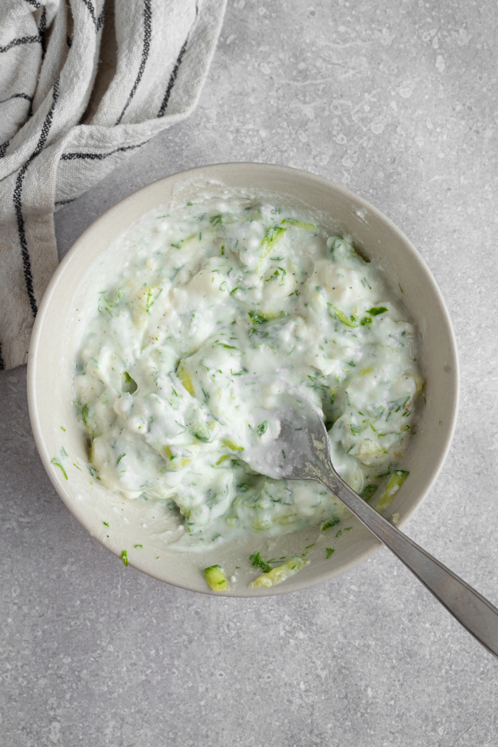 A small bowl with a spoon mixing tzatiki sauce. The bowl is on a white counter with a white striped napkin.