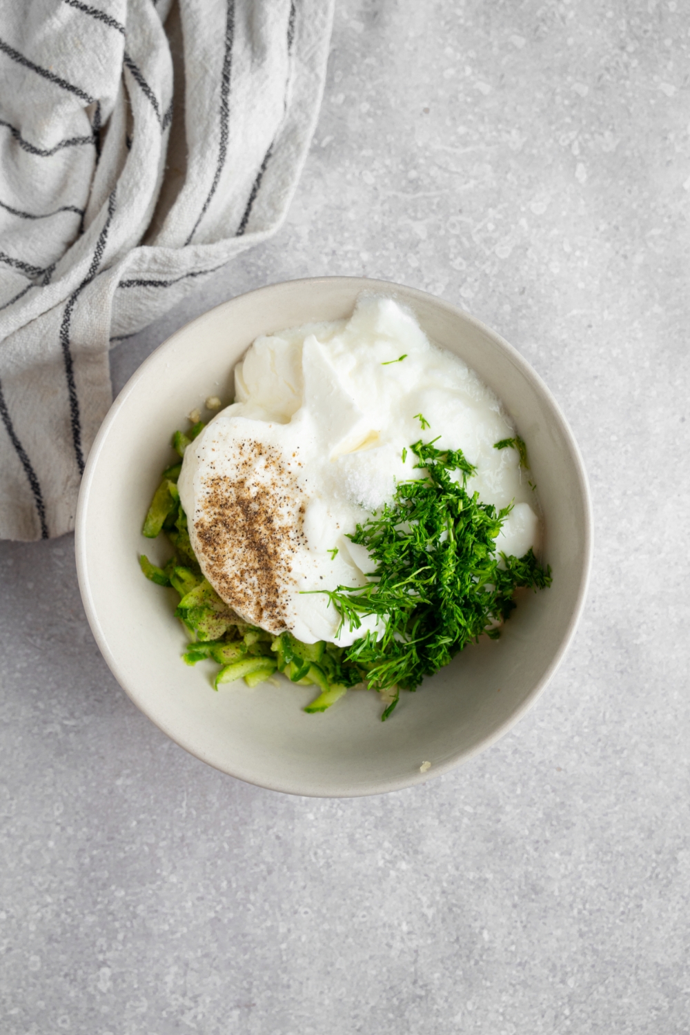 A small bowl with ingredients to make tzatziki incluing greek yogurt, dill, and cucumber. The bowl is on a white counter with a white striped napkin.