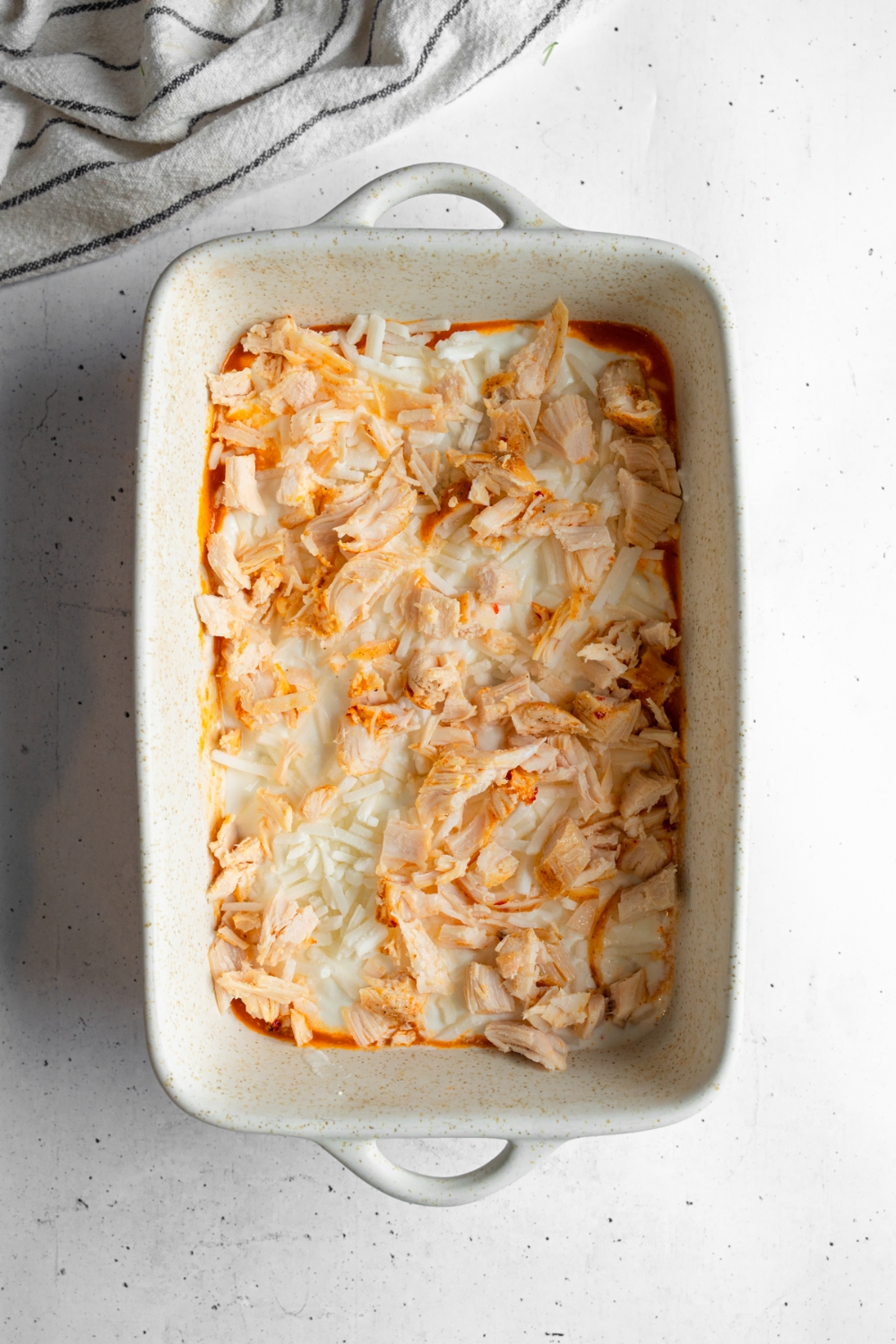 A baking dish with rotisserie chicken lasagna with mozzarella cheese and rotisserie chicken layered over noodles and sauce. The dish is on a white counter with a white striped napkin.