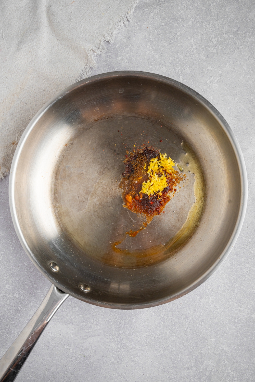 A stainless steel skillet with olive oil and seasonings. The skillet is on a white counter with a white cloth napkin.